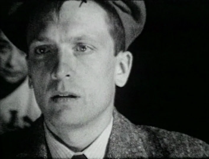 Cowards Bend The Knee (2003) movie still: closeup — A dimly lit indoor space with a dark background. A man in a cap gazes ahead.
