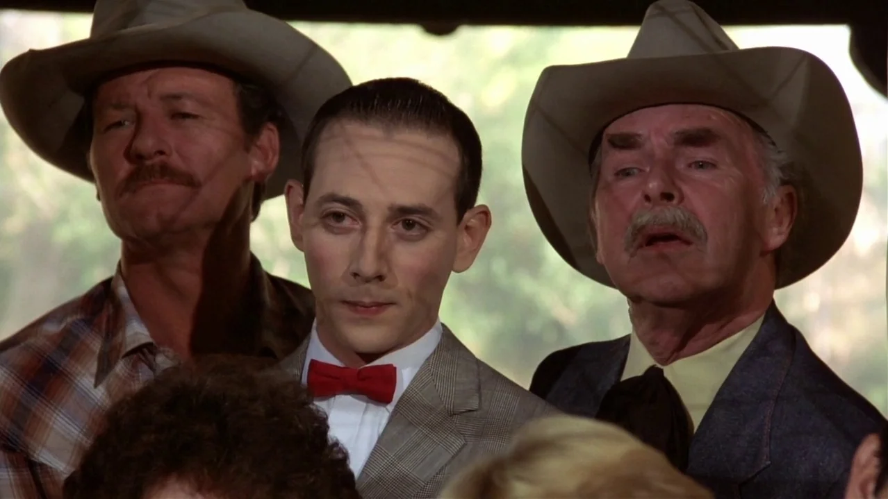 Pee-Wee’s Big Adventure (1985) movie still: medium — Three men stand side-by-side in an outdoor setting beneath a dark structure. Each man wears a…