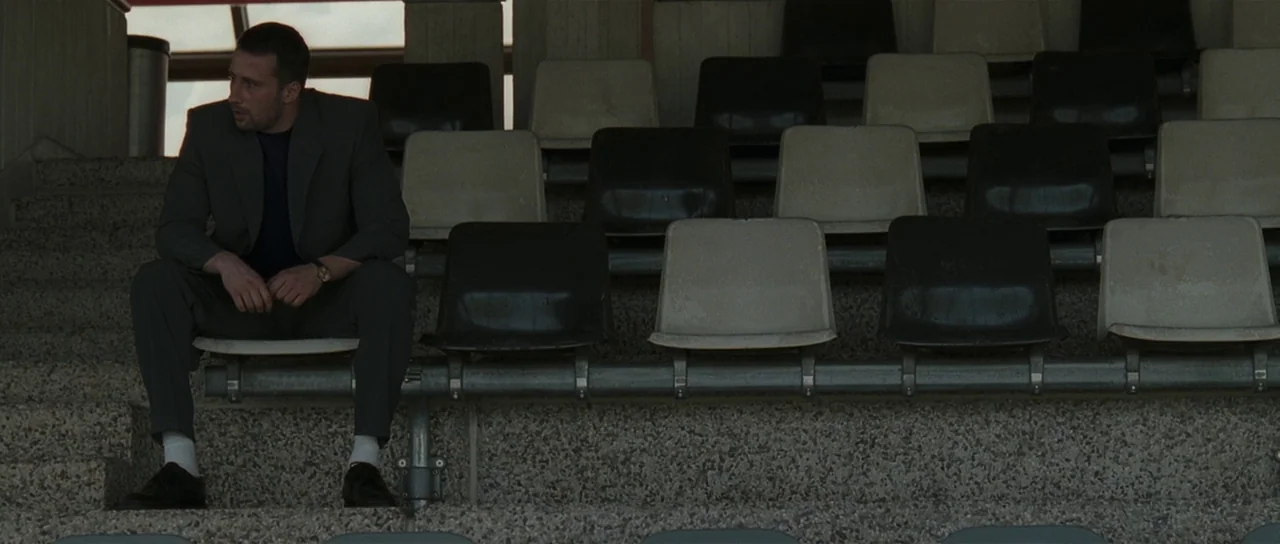 Bullhead (2011) movie still: medium — A man sits alone on empty stadium seating featuring rows of black and white chairs. The structure is…
