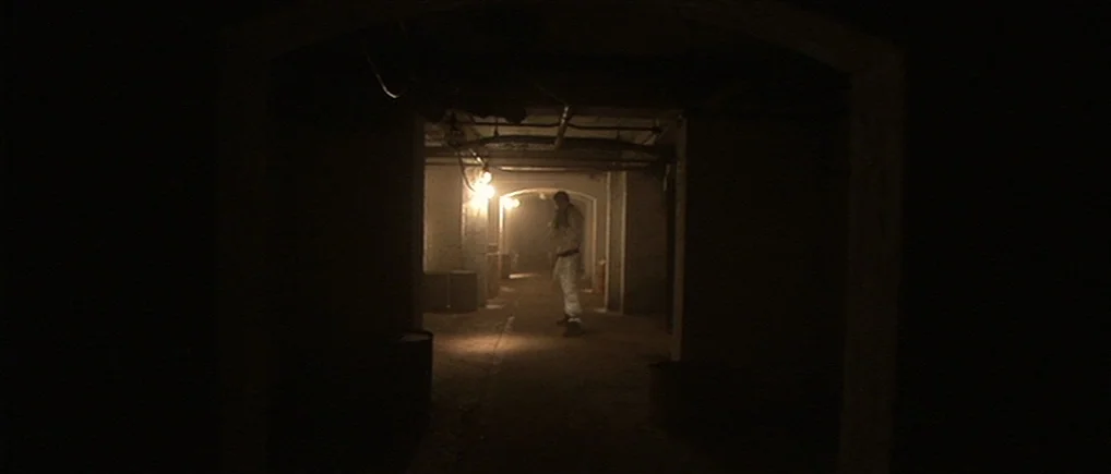 Session 9 (2001) movie still: wide — A dimly lit industrial basement features concrete archways and exposed ceiling pipes. A lone figure in white…