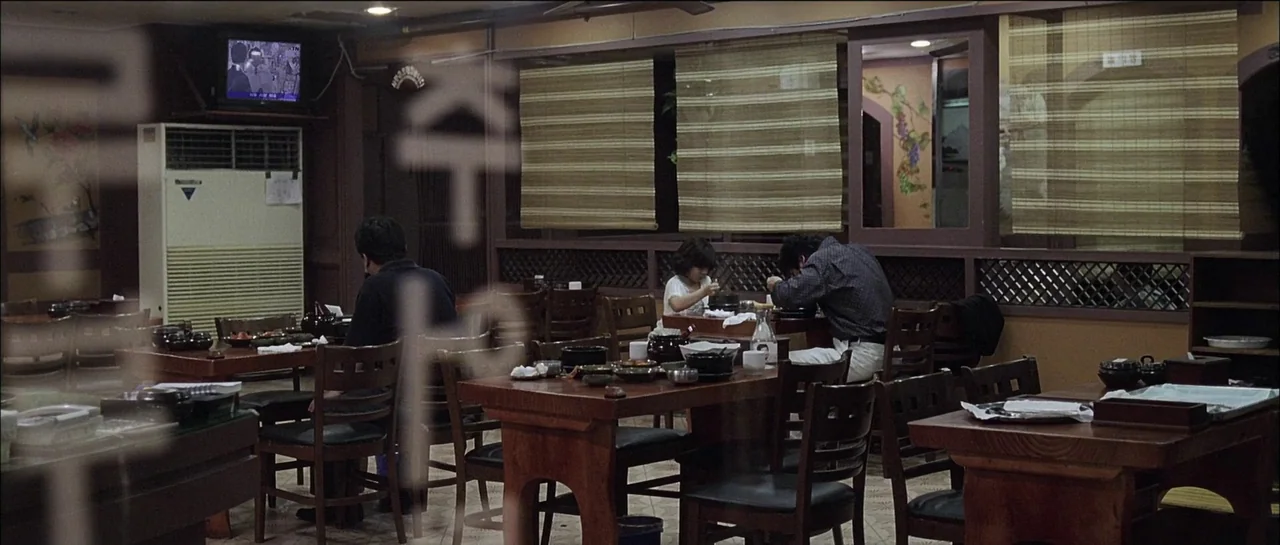 The Chaser (2008) movie still: wide — A quiet restaurant interior features wooden tables and bamboo window shades at night. Three individuals are…