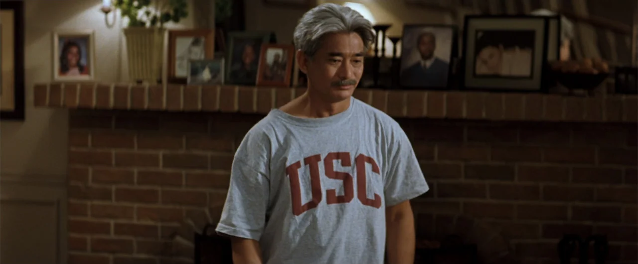 Lethal Weapon 4 (1998) movie still: medium — A domestic interior features a man standing before a brick fireplace mantle adorned with framed…