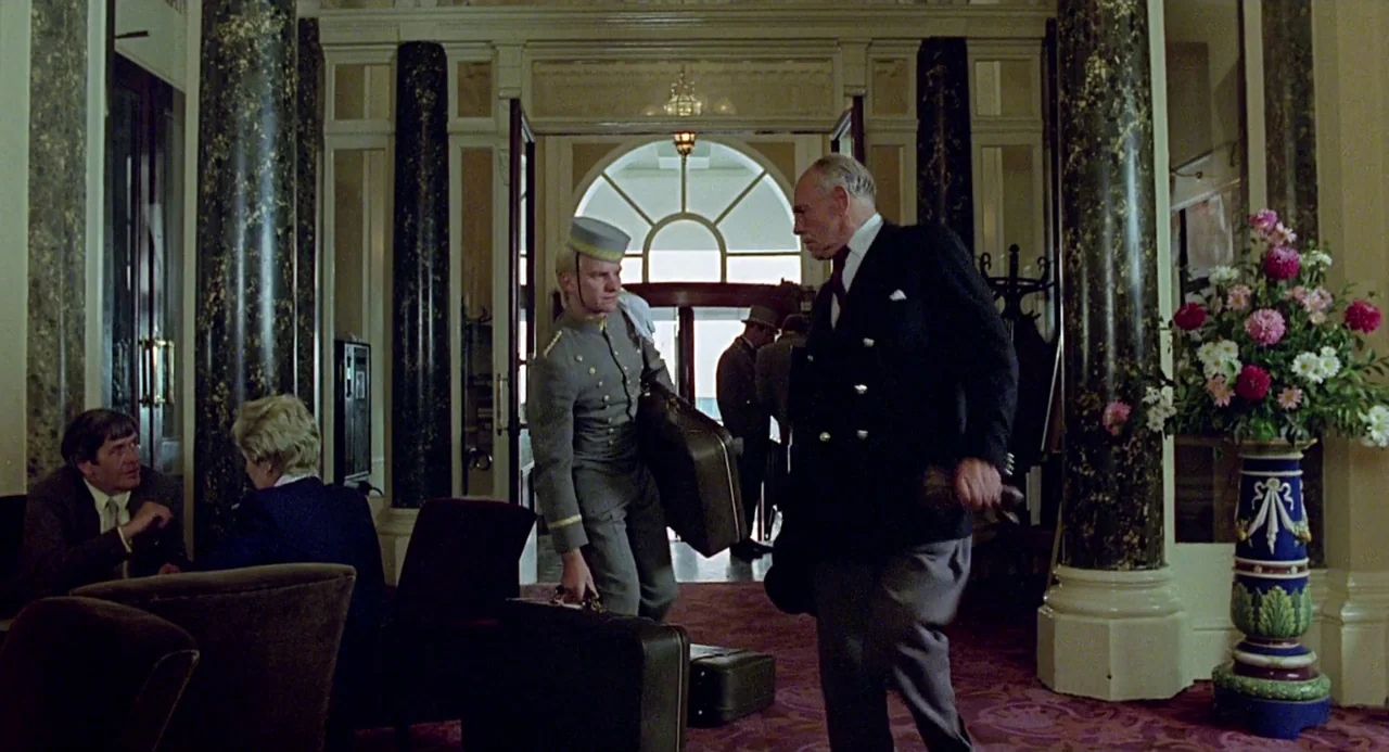 Quadrophenia (1979) movie still: fullbody — A classic hotel lobby features marble pillars and patterned carpeting near an open entrance. A bellboy…