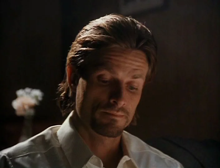 Head of the Family (1996) movie still: closeup — A man stands in a dimly lit, dark-toned interior space with blurred flowers behind him. He gazes…