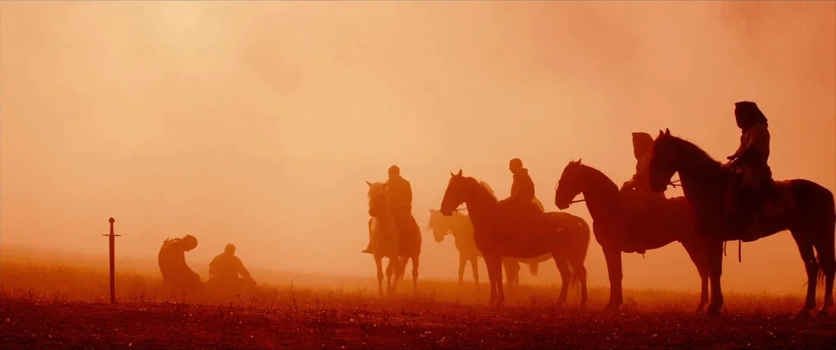 Macbeth (2015) movie still: wide — A vast, dusty field bathed in intense orange light under a low sun. Several riders on horseback stand near two…