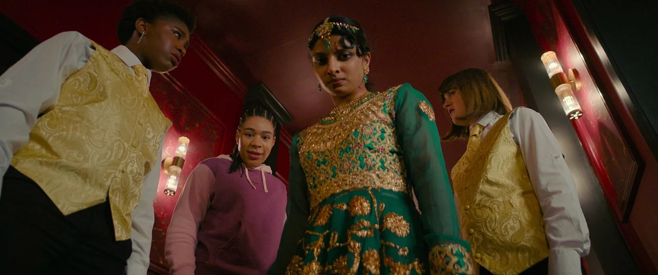 Polite Society (2023) movie still: medium — A narrow dimly lit corridor with deep red walls and decorative gold molding. Four young women stand in a…