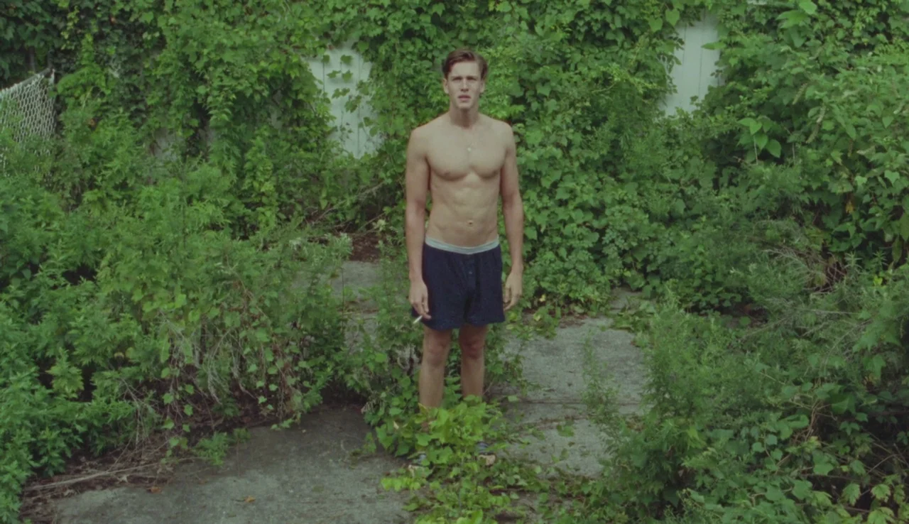 Beach Rats (2017) movie still: fullbody — A dense overgrown backyard features lush green foliage surrounding a concrete paved path. A man in boxer…