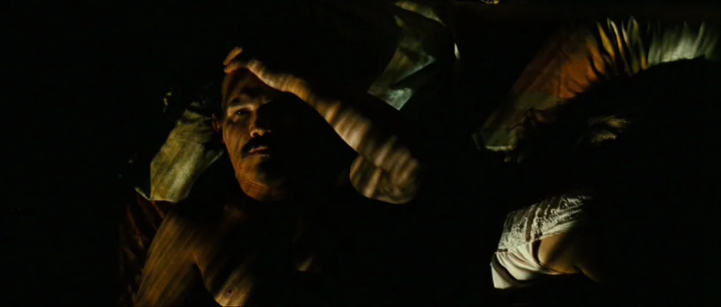 No Country For Old Men movie still: medium — A dark, dimly lit bedroom at night with a man and woman in bed. The man lies awake while the woman…