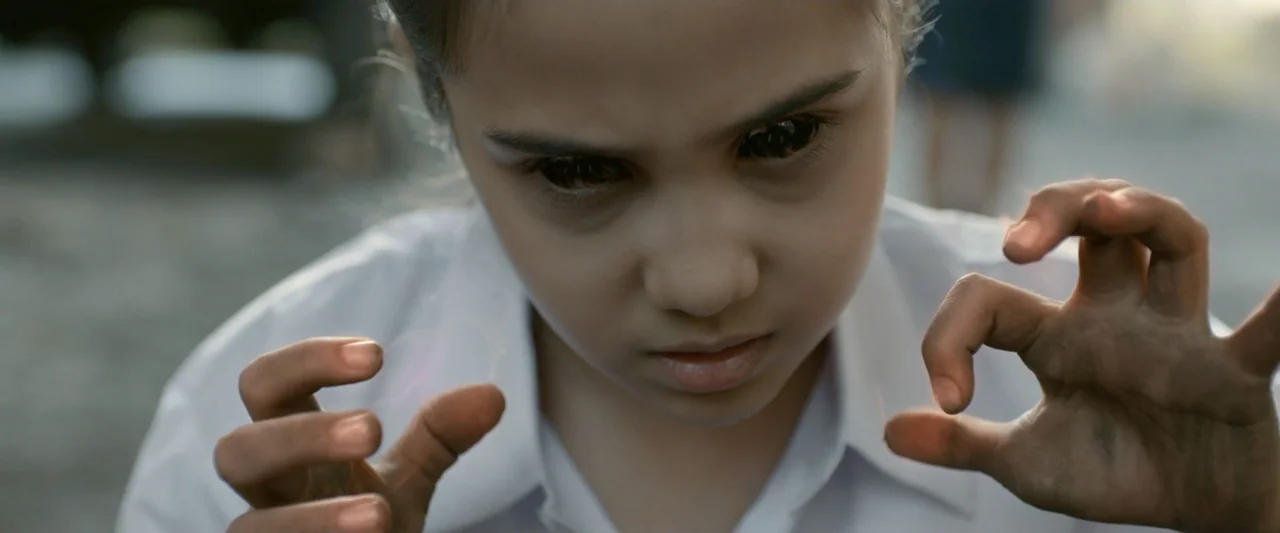 The Chanting 3 (2008) movie still: closeup — A young girl stands in an outdoor area with blurred background elements. She holds her hands out in…