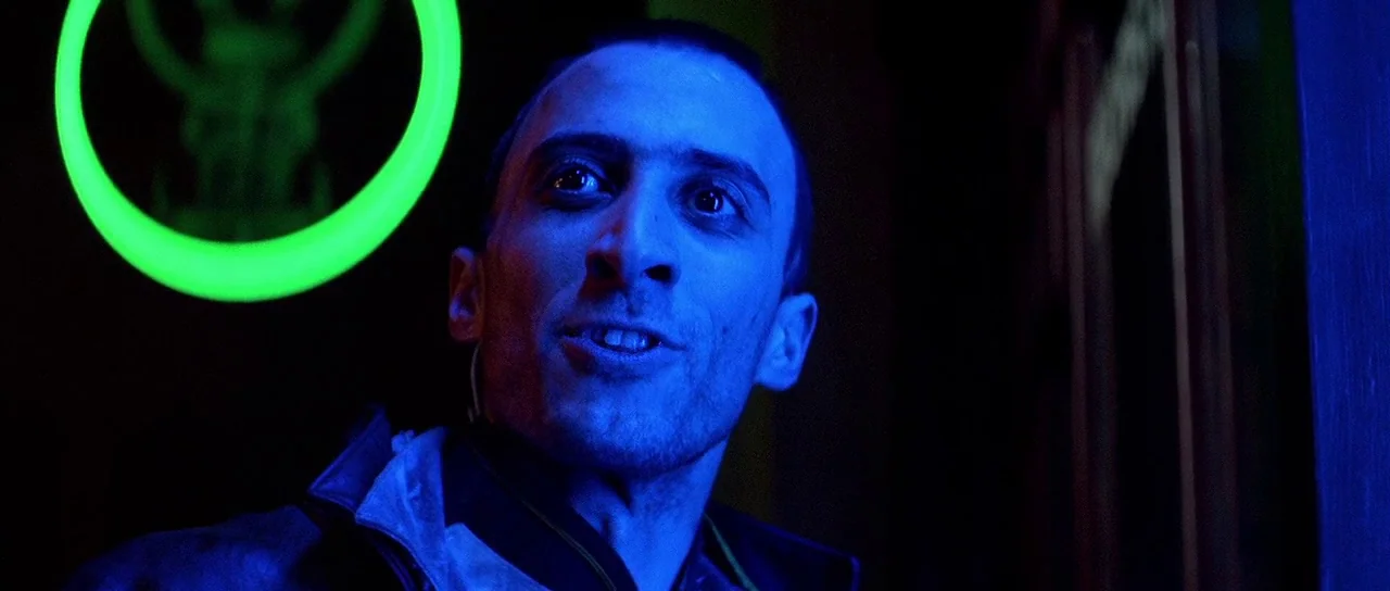 Nikita (1990) movie still: closeup — A dark space lit with intense blue and neon green colored lights. A man with short hair looks off to the side…