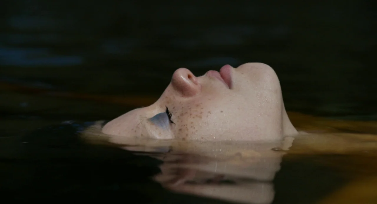 X (2022) movie still: closeup — A person floats in dark, still water with only their profile visible above the surface. The head is partially…