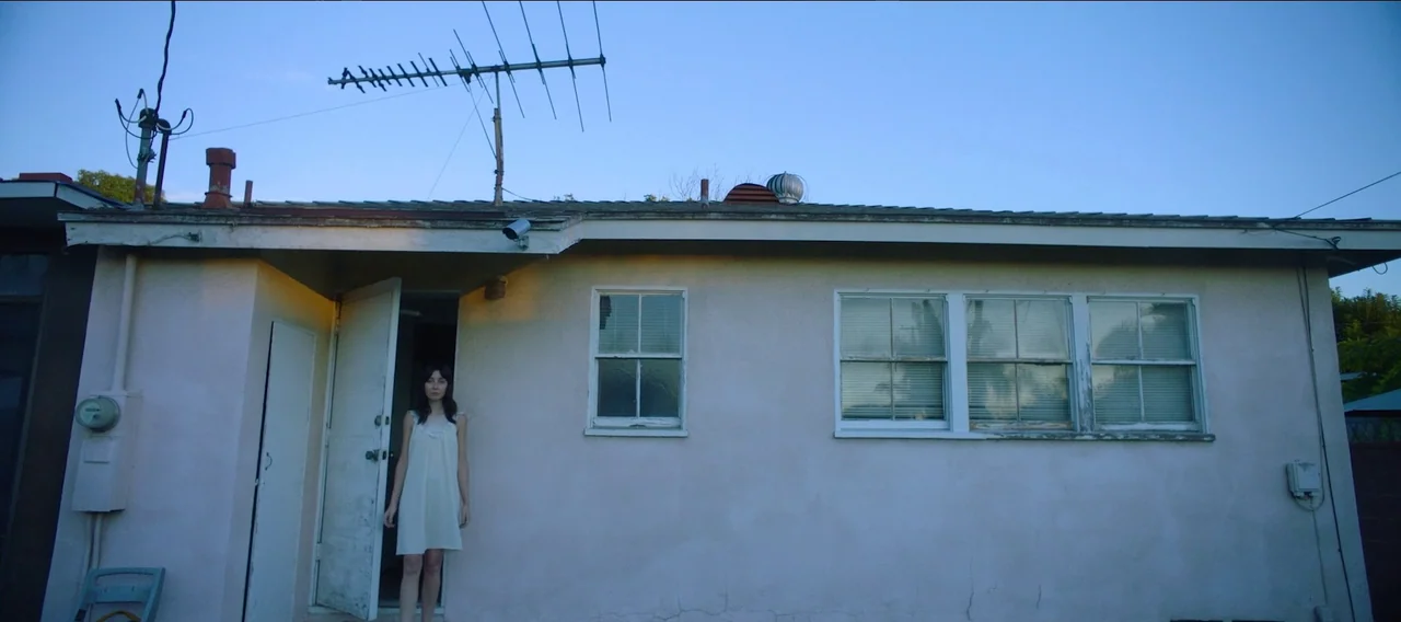 Fuzzy Head (2023) movie still: wide — The exterior of a pale suburban building features a television antenna and windows. A woman in a white dress…