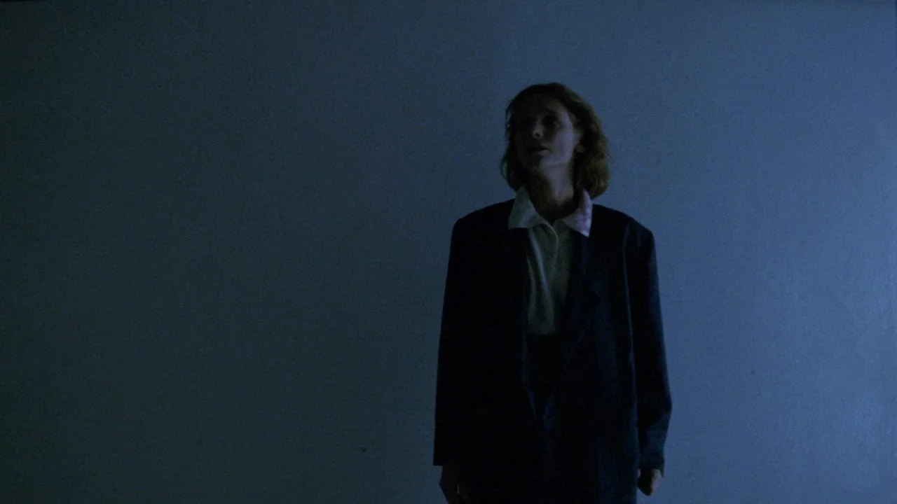 The Sect (1991) movie still: medium — A person stands in a dimly lit room with plain, solid-colored walls. The subject is positioned slightly…