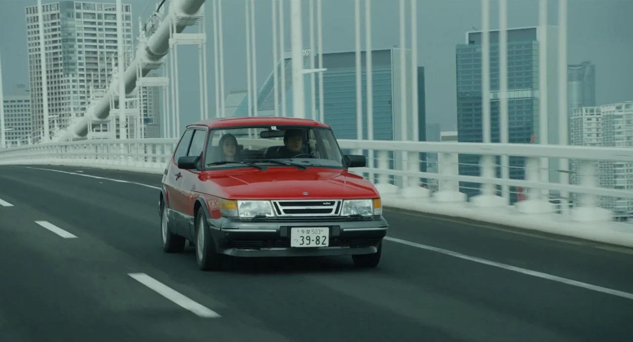 Drive My Car (2021) movie still: wide — A red Saab hatchback drives across a modern white suspension bridge during an overcast day. Two people…