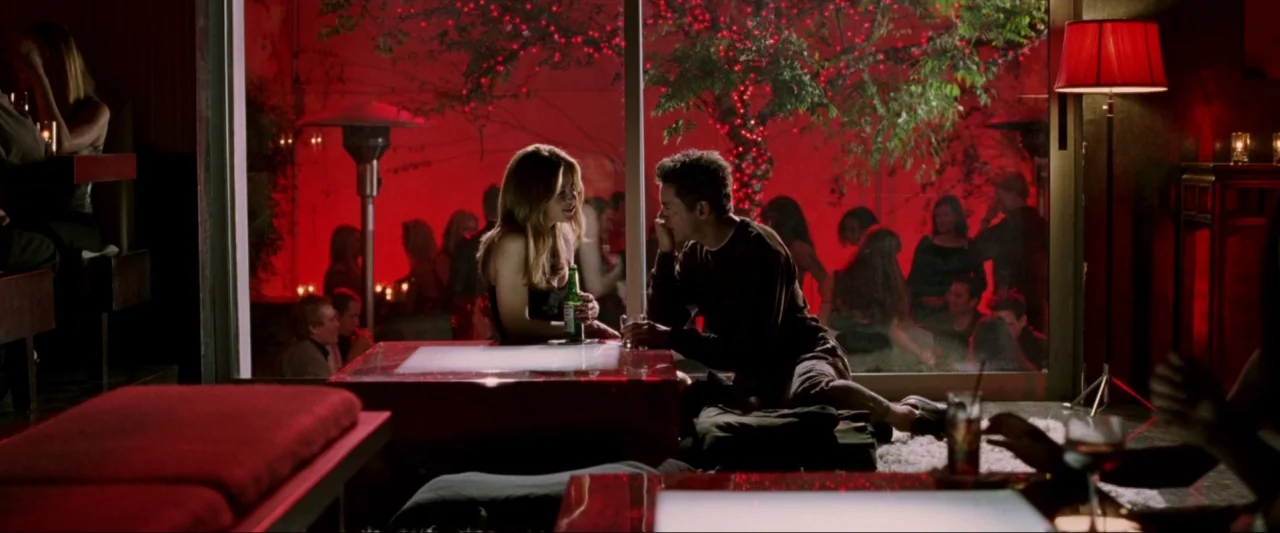 Kiss Kiss Bang Bang (2005) movie still: medium — A dark lounge features a large glass wall looking out onto a red-lit terrace. Two people sit at a…