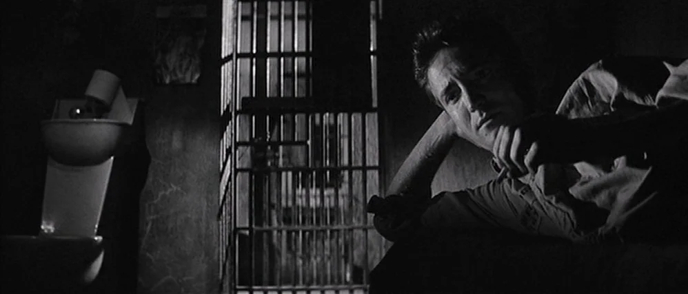 In Cold Blood (1967) movie still: medium — A dimly lit room with iron bars separating the sleeping area from the corridor. A man lies on a cot with…