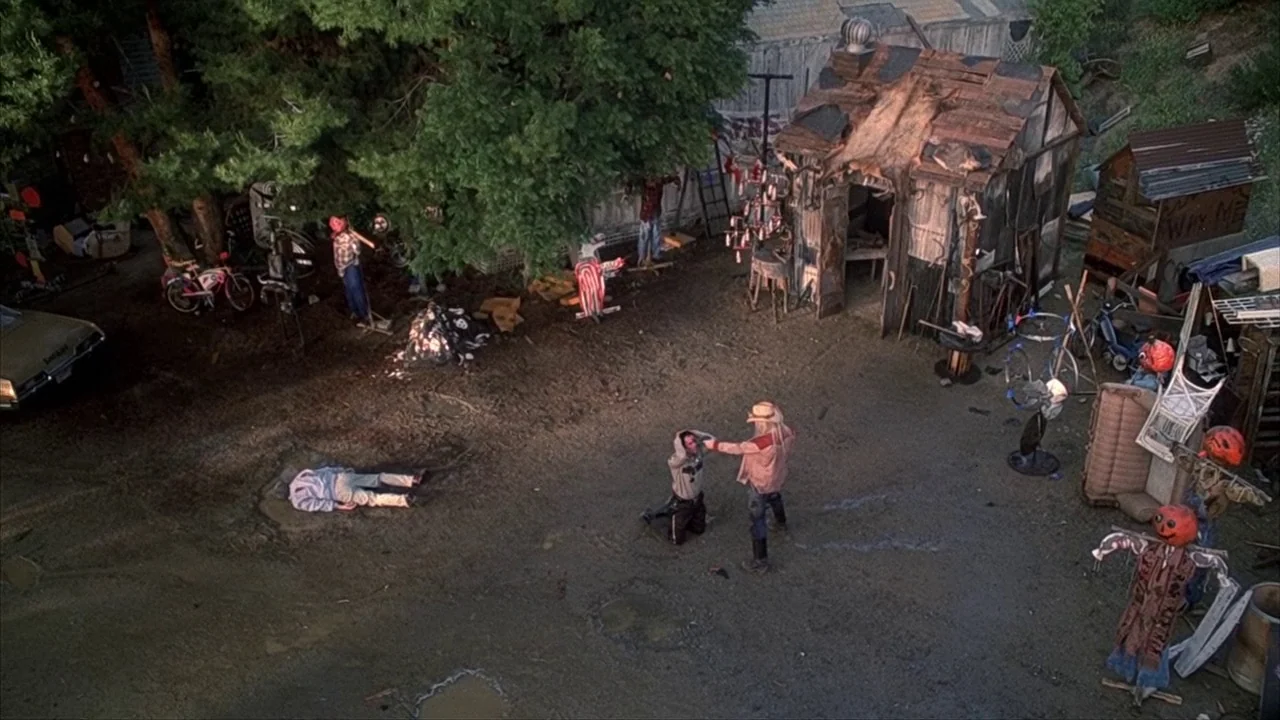 House of 1000 Corpses (2003) movie still: wide — A cluttered outdoor lot contains a small shack, old bicycles, and assorted debris. A man in a…