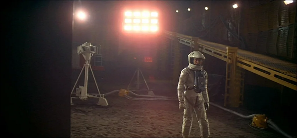 2001: A Space Odyssey (1968) movie still: fullbody — A dark, sandy lunar landscape features a large floodlight and a yellow elevated ramp. An…