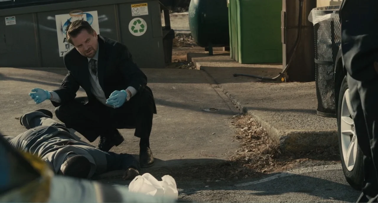 Lila & Eve (2015) movie still: medium — A paved area features a large metal dumpster and a trash bin nearby. A man in a suit crouches to examine a…