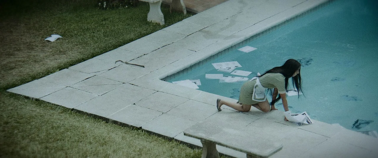 La Llorona (2019) movie still: wide — A stone-paved pool deck surrounds a turquoise swimming pool filled with scattered white papers. A young woman…