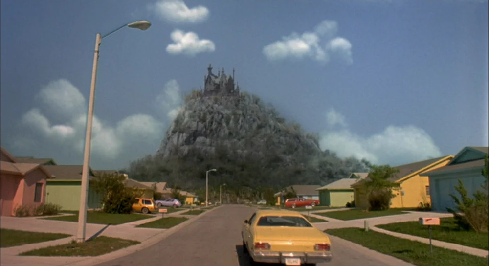 Edward Scissorhands (1990) movie still: establishing — A suburban street lined with brightly colored houses leads to a large, rocky mountain. A…