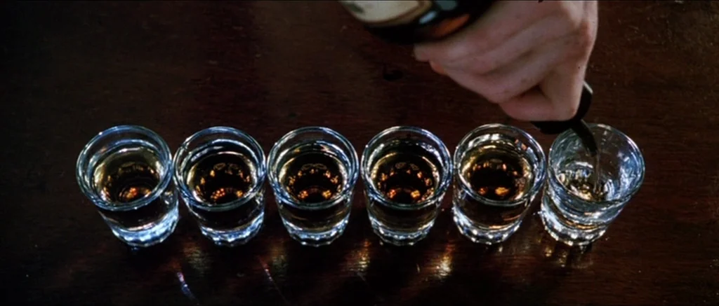 Death Proof movie still: closeup — A dark wooden surface holds a neat row of six small crystal shot glasses. A hand tilts a dark glass bottle to…