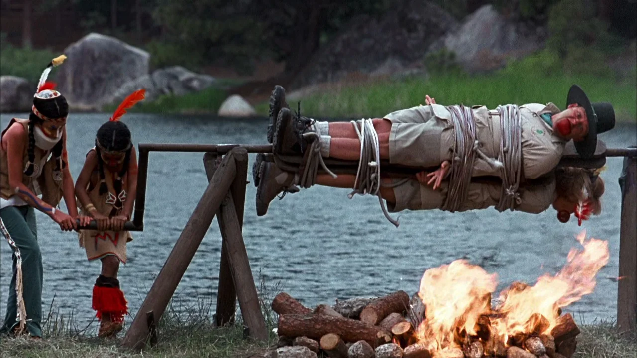 Addams Family Values (1993) movie still: wide — A lakeshore scene features a spit rotating over a campfire near large boulders. Two people in…