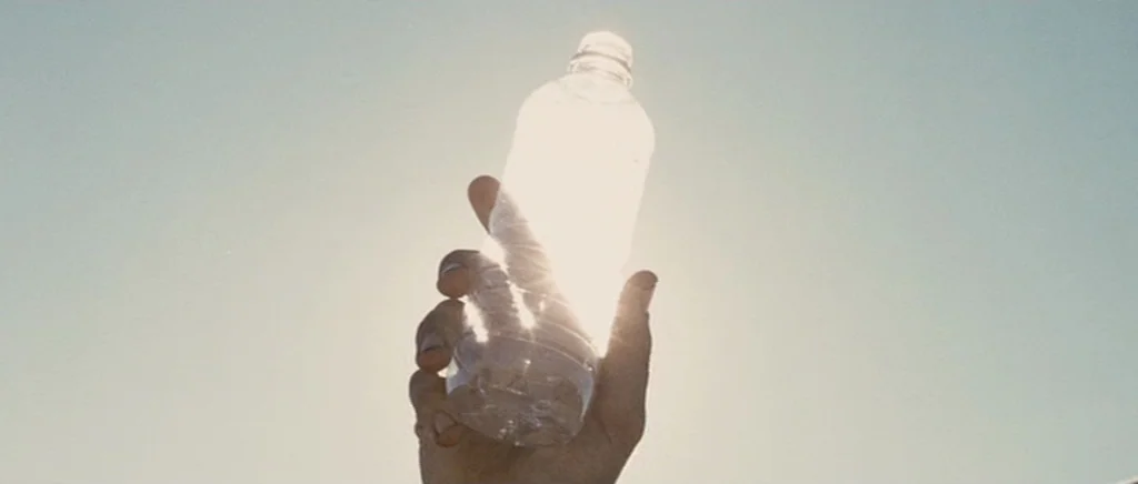 Jarhead movie still: closeup — A clear plastic bottle is held up against a bright, featureless sky. The sun shines directly behind the bottle,…