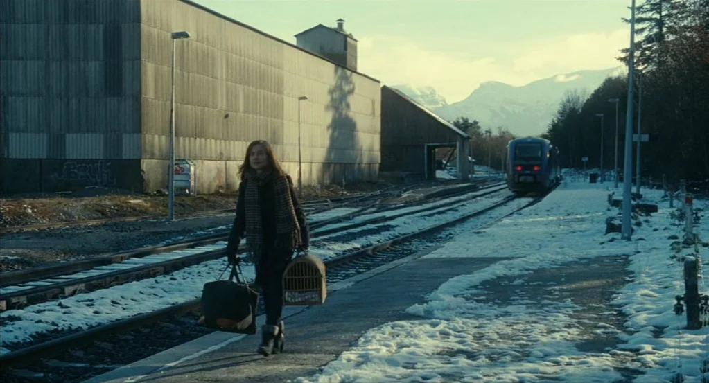 Things to Come (2016) movie still: fullbody — A woman walks along a snowy train platform next to a long metal shed. In the distance a train…