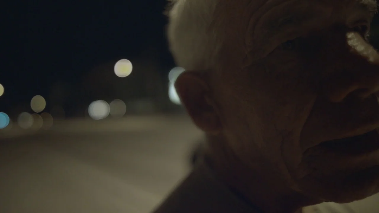 Florida Man (2015) movie still: closeup — An elderly man is framed in close-up against a dark out-of-focus background. The background features…