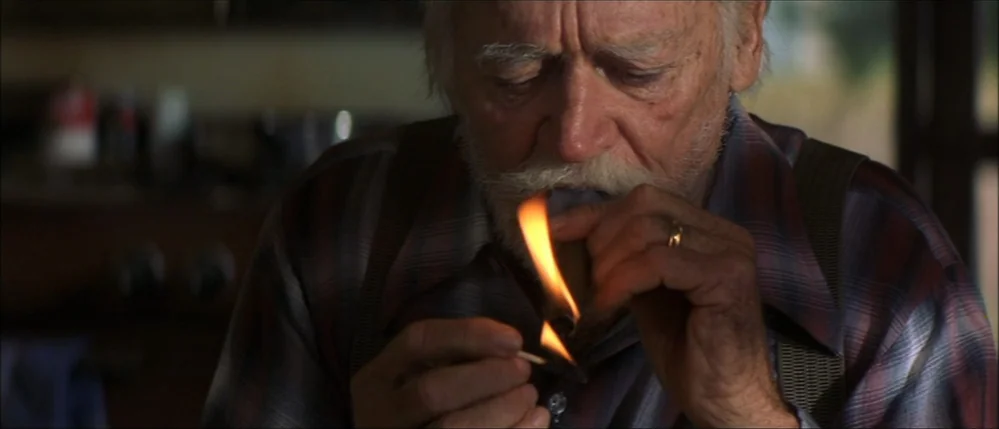 The Straight Story (1999) movie still: closeup — An elderly man is shown in close up view in a darkened interior. He holds a burning match to light…