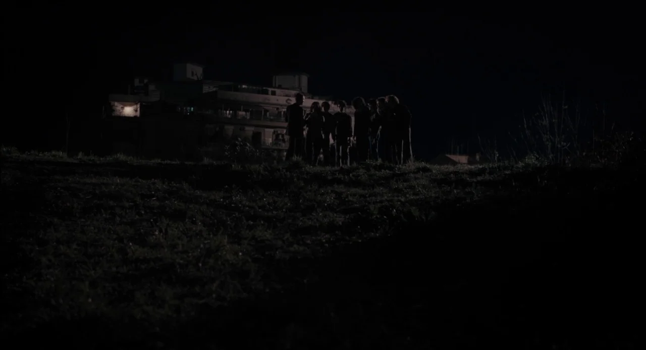 Pasolini (2014) movie still: wide — A dark, grassy field stretches out at night with a distant building silhouette visible. A group of several…