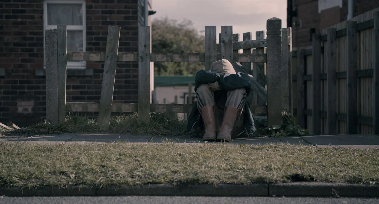 The Selfish Giant (2013) movie still: medium — A suburban sidewalk is bordered by a weathered wooden picket fence and brick architecture. A single…