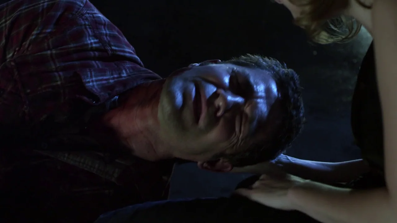 Hellraiser: Revelations movie still: closeup — A man lies on a dark floor in a dimly lit space. A woman reaches out to touch his head with her hand.
