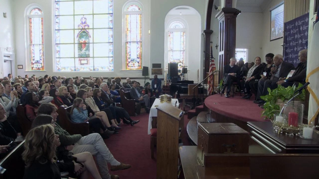 The Soul of America (2020) movie still: wide — A church interior with wooden pews and large stained glass windows seating a crowd. Several…