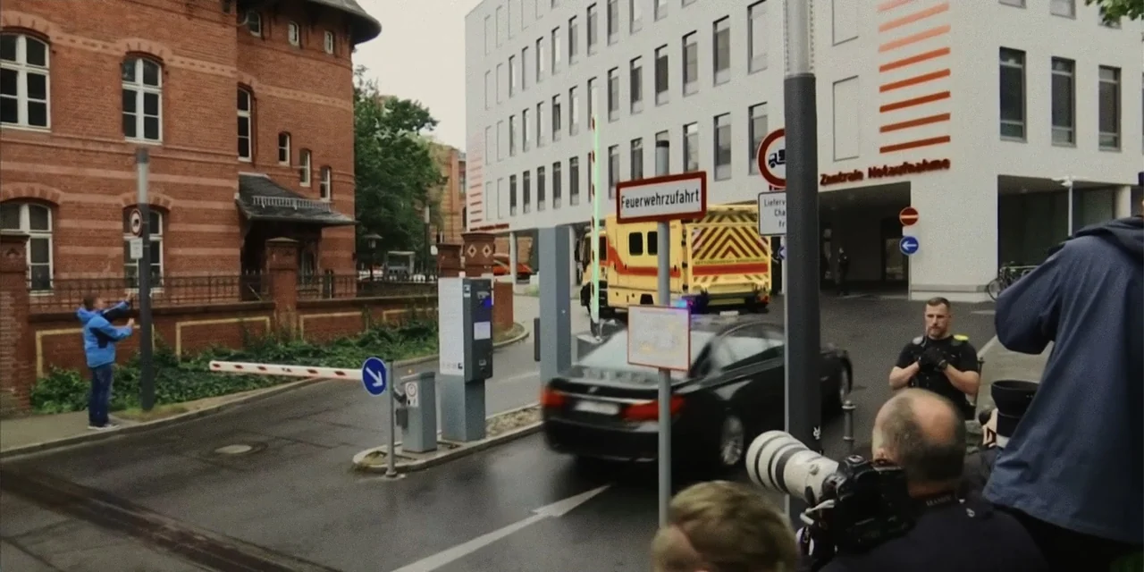 Navalny (2022) movie still: wide — A modern hospital exterior features a gated entrance with a security barrier and signage. A police officer stands…