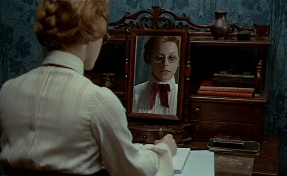 Two English Girls (1971) movie still: medium — A woman sits at a wooden vanity with dark blue floral wallpaper in the background. She is writing in…