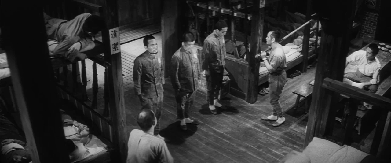 The Human Condition II: Road to Eternity (1959) movie still: wide — A wooden bunkroom with tiered beds, rifles leaning against posts, and dark…