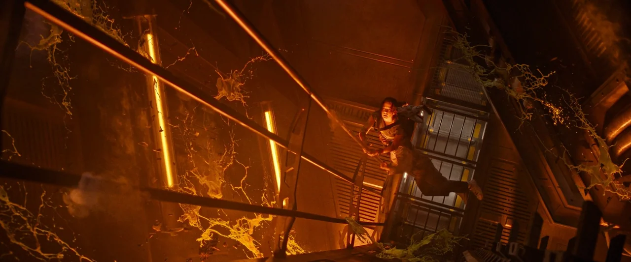 Alien: Romulus (2024) movie still: wide — A high-tech industrial hallway with metal grates and yellow neon accents. A young woman grips a rail while…