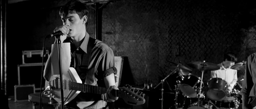 Control movie still: medium — A dark, industrial rehearsal space features concrete walls and exposed wiring in the background. A young man sings…