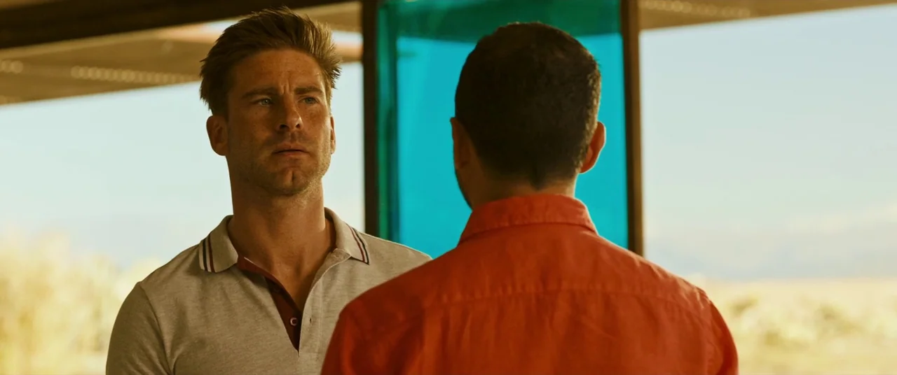Revenge (2017) movie still: medium — A modern building with expansive glass windows overlooks a dry, arid landscape. One man faces another man who…