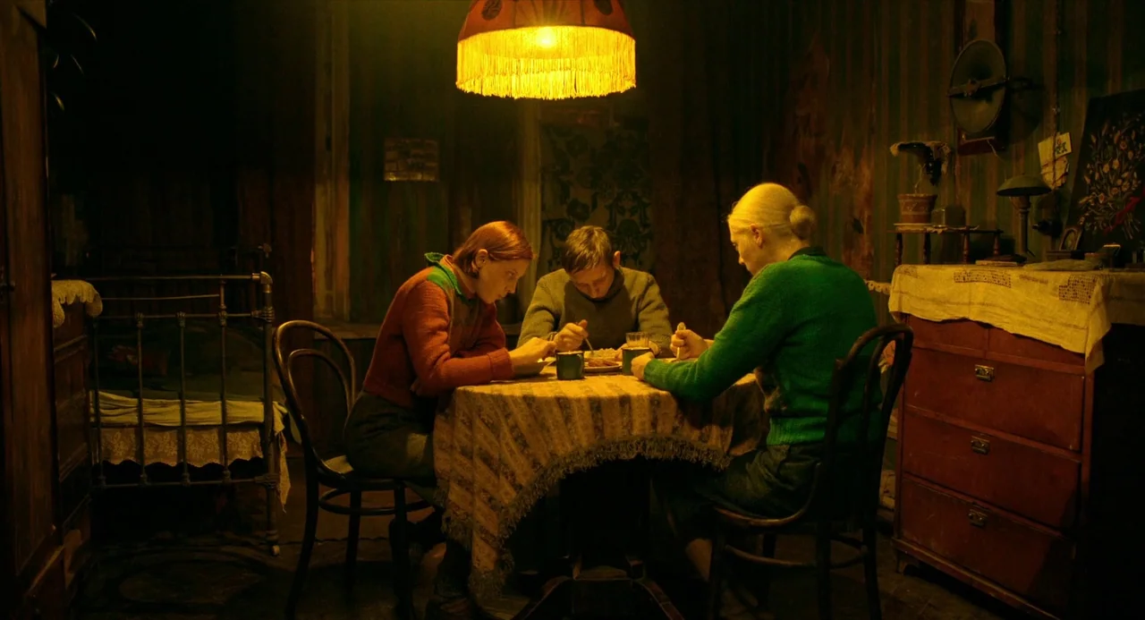 Beanpole (2019) movie still: medium — A dim room features a small table with three people eating under a lamp. The walls have textured, dark green…