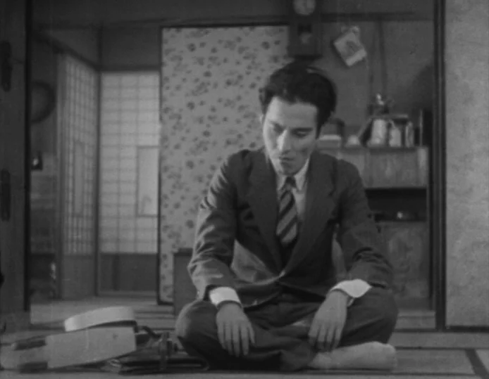 Tokyo Chorus (1931) movie still: medium — A traditional Japanese room features straw tatami mats and paper-paneled sliding doors. A young man sits…