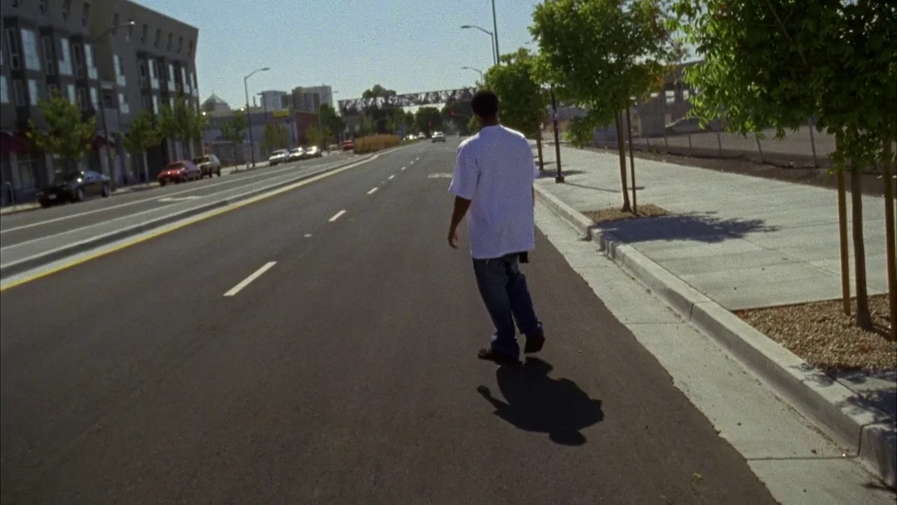 Fruitvale Station (2013) movie still: fullbody — A wide, multi-lane urban road stretches into the distance under a clear blue sky. A young man walks…
