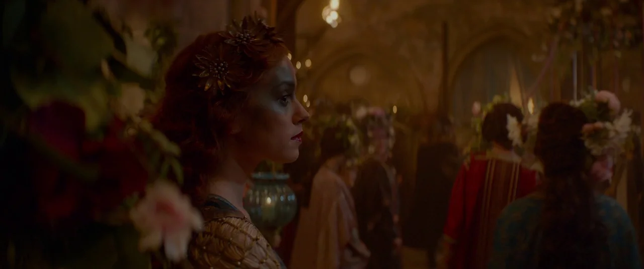 Ophelia (2018) movie still: medium — A dimly lit hall decorated with flowers is occupied by a gathering of people. A woman with an ornate floral…