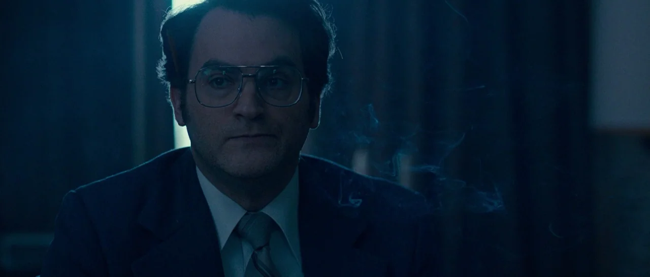 Pawn Sacrifice (2014) movie still: medium — A man sits in a dark, dimly lit environment with blue hues. He has a pencil tucked behind his ear and…