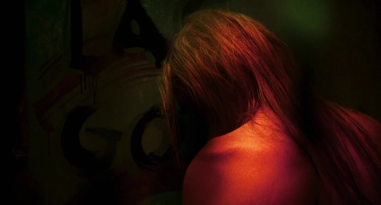 3 From Hell (2019) movie still: closeup — A dimly lit space features a wall covered with dark, layered spray paint markings. A woman with long hair…