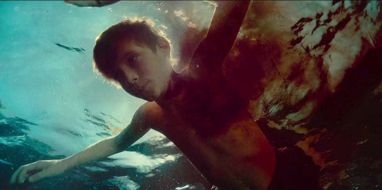 Mute (2018) movie still: medium — A teenage boy swims through calm, translucent blue water in a natural setting. The camera captures him from below…