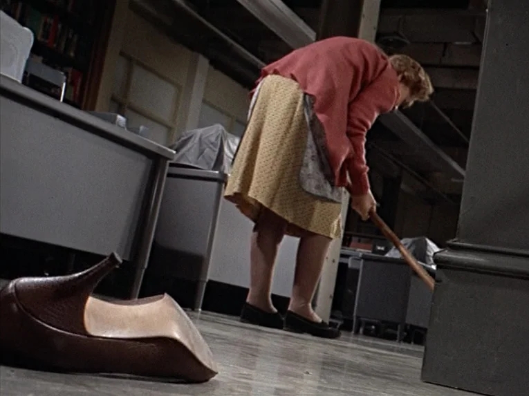 Marnie (1964) movie still: medium — A cluttered, dimly lit storage basement with gray metal desks and filing equipment. An elderly woman in a pink…