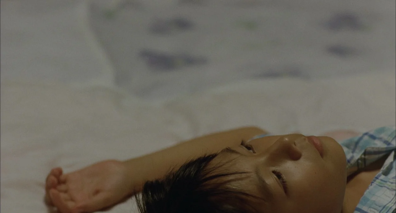 Still Walking (2008) movie still: closeup — A young child lies on a white bed with blurred fabric textures behind. The child rests their head on the…